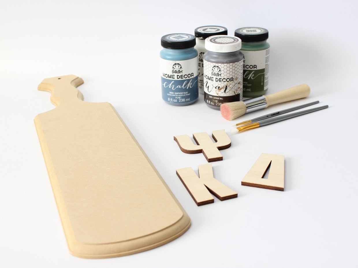 A blank wooden Greek paddle beside cut wooden Greek letters and craft paint